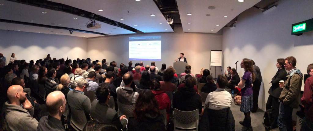 A room where a research presentation is happening. Daniel Trielli is speaking in front of a lectern, showing slides projected on a screen.