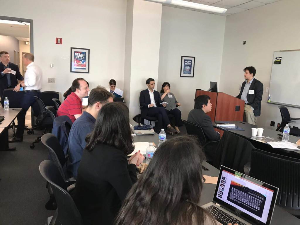 A room full of people watching research presentations. Daniel Trielli is behind a lectern, speaking.