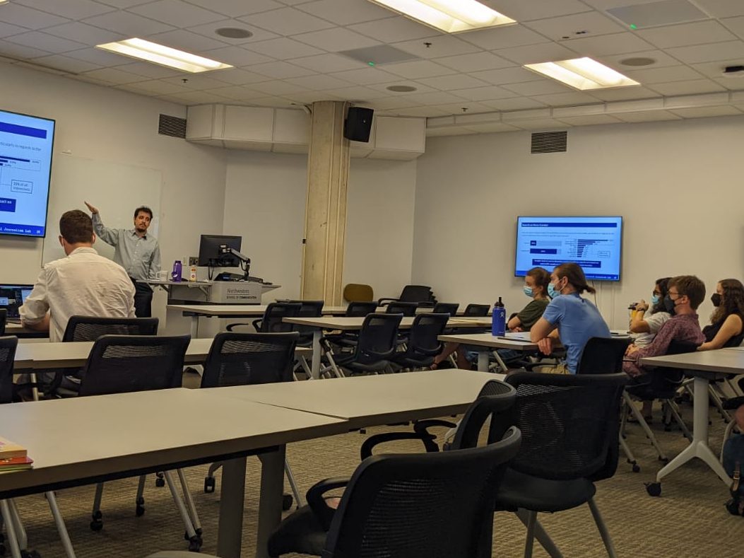 Daniel Trielli presenting slides shown in large displays, talking to a room with people sitting and watching.